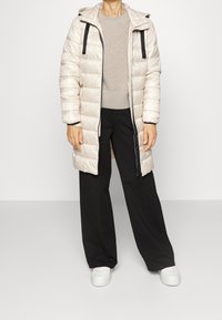 Cappotto imbottito beige chiaro con chiusura a zip, dettagli neri e cappuccio, indossato sopra un maglione chiaro e pantaloni neri larghi. Sneakers bianche ai piedi.