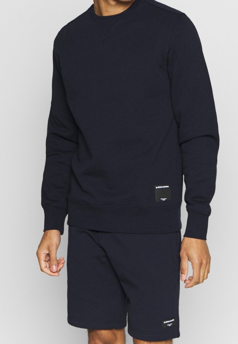 Sweat-shirt bleu marine avec un col ras du cou, des poignets et un ourlet côtelés. Présente un petit écusson logo en bas à gauche. Confectionné en matériau doux.