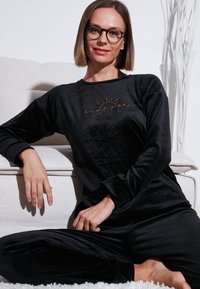 Zwart fluwelen loungewear set met lange mouwen, voorzien van een gouden geborduurd logo. Getoond tegen een lichtgekleurde bank.