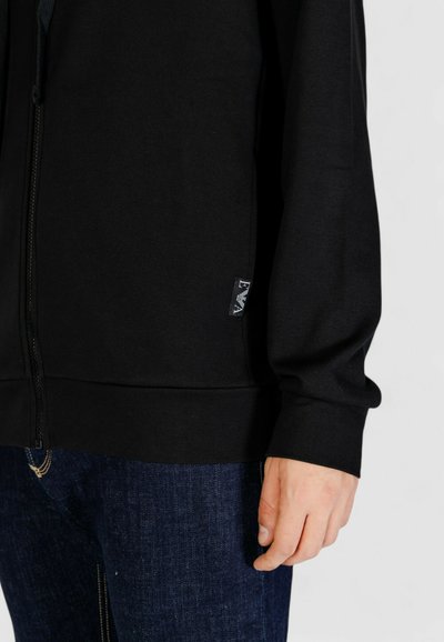Felpa nera con zip realizzata in un tessuto texturizzato, con polsini e orlo a costine. Presenta un'etichetta del marchio sulla parte inferiore sinistra. Abbinata a jeans blu.