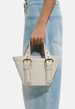 Ręka trzyma małą białą torebkę z złotymi klamrami na paskach, noszoną z jasnoniebieskimi dżinsami.