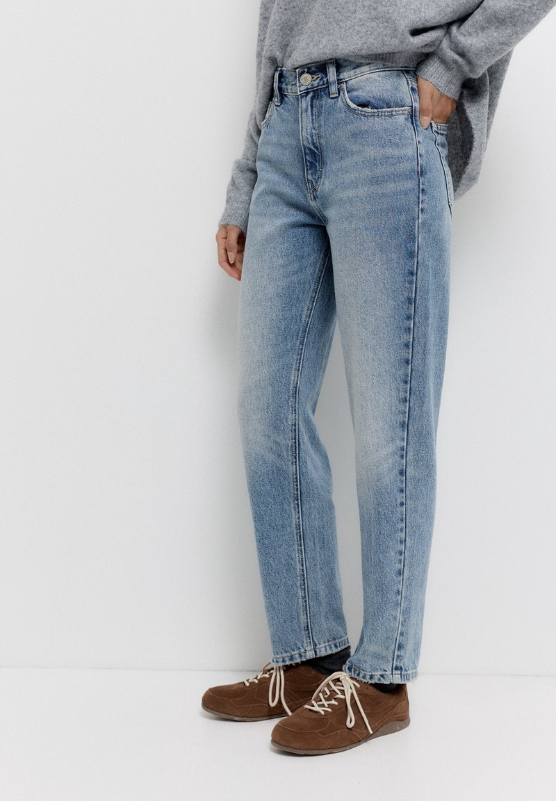 Personne portant un jean droit bleu clair, un pull gris et des chaussures marron à lacets, debout avec une main dans la poche.