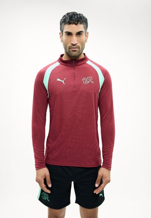 Άντρας που στέκεται προς τα εμπρός, φορώντας μακρυμάνικο μπορντό μπλουζάκι Puma με λογότυπο και μαύρο σορτς με ταιριαστά ανοιχτό μπλε διακοσμητικά στοιχεία.