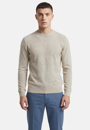 Ung mand iført en lysebeige tekstureret sweater og blå skræddersyede bukser, der står mod en ensfarvet hvid baggrund.