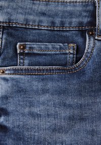 Vaqueros de mezclilla con una textura desgastada, que cuentan con un pequeño bolsillo frontal, adornos de metal latonado y costuras amarillas contrastantes sobre una tela azul.