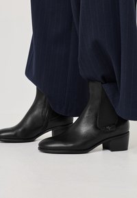 Botas de cuero negras hasta el tobillo con paneles laterales elásticos, puntera redonda, tacón bajo y detalles discretos de la marca en el lado exterior.