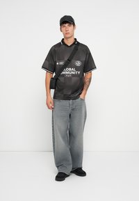 Czarny graficzny t-shirt z białymi akcentami, duże jasnoszare dżinsy, czarne buty typu slip-on i czapka w kolorze czarnym. Prosta czarna torba na ramię.