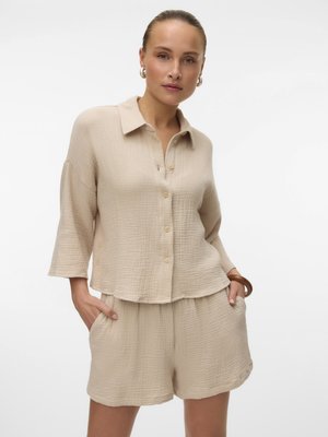 Kvinde med glat hår, iført beige textured skjorte med knapper og matchende shorts, hænderne i lommerne, poserer mod en hvid baggrund.