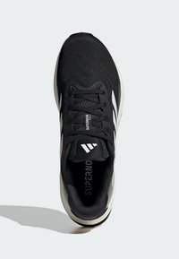 Chaussure de sport noire avec une tige en mesh texturé, accents blancs et une semelle rembourrée. Dispose d'un système de laçage central et d'un logo sur la languette.