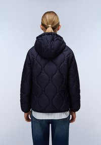 Marinblå quiltad pufferjacka med huva, med en texturerad mönstring och elastiska ärmar, bärs över en ljusrandig skjorta och jeans.