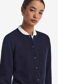 Marineblauer Cardigan aus weichem Stoff mit rundem Ausschnitt, Knopfverschluss und glatter Textur. Wird über einem weißen Hemd mit Kragen getragen.