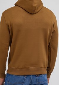 Ruskea huppari joustavasta kankaasta, jossa on kengurun tasku, resorit hihansuissa ja helmassa. Takapuoli on yksivärinen ja pehmeäpintainen.