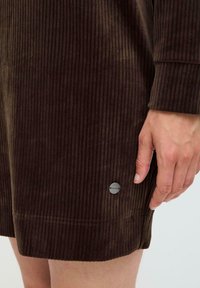 Robe en velours côtelé marron avec des rayures verticales, doté d'un ourlet arrondi et d'un accent logo en métal argenté près du bas. Texture lisse.