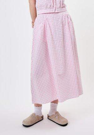 Vrouw met een roze-witte geruite rok, witte sokken en beige instapschoenen tegen een effen witte achtergrond.