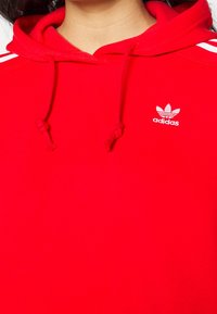 Röd Adidas hoodie med dragsko i huvan, vita kontrastlinjer på axlarna och vit logotyp framtill. Mjuk tygtextur.