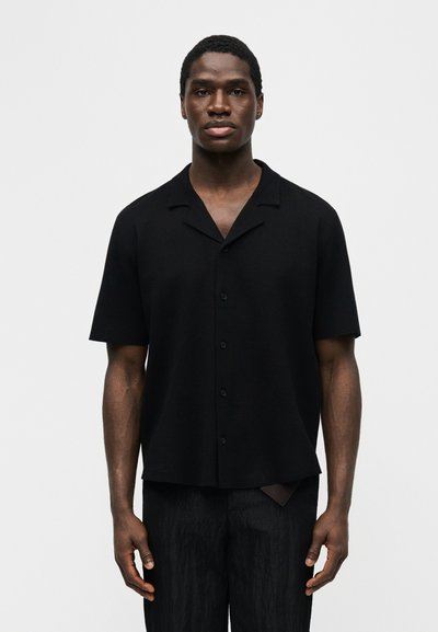 Άνδρας που στέκεται με το σώμα στραμμένο προς τα εμπρός, φορώντας μαύρο κοντομάνικο πουκάμισο με κουμπιά και μαύρο παντελόνι, μπροστά σε απλό λευκό φόντο.