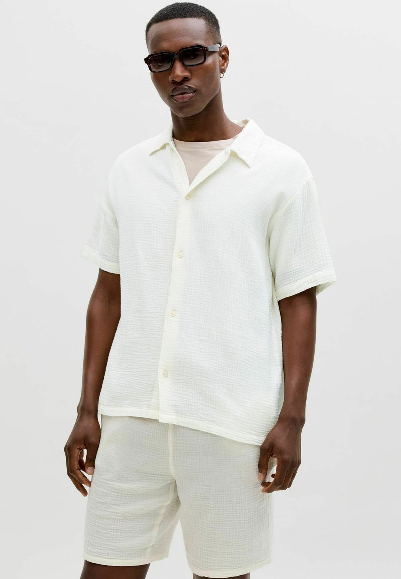 Homme portant une chemise boutonnée à manches courtes texturée de couleur blanc cassé et un short assorti, accessoirisé avec des lunettes de soleil foncées et une petite boucle d'oreille.
