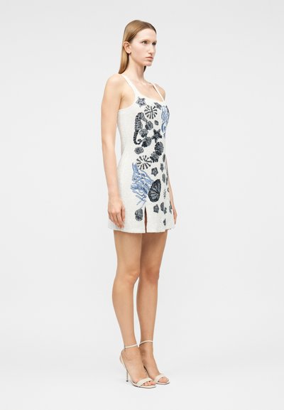 Γυναίκα που φοράει λευκό μίνι φόρεμα χωρίς μανίκια με μπλε και μαύρα σχέδια κοχυλιών, σε συνδυασμό με λευκά πέδιλα με ψηλό τακούνι, όρθια πλάγια.