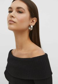 Schwarzes, geripptes Off-Shoulder-Top mit weitem Ausschnitt. Große silberne Creolen mit polierter Oberfläche ergänzen den Look. Glatte Hautfarbe.