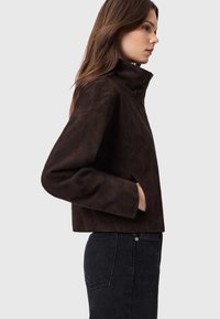 Giacca in camoscio marrone con colletto alto e design corto. Presenta tasche laterali e una texture liscia, abbinata a jeans di denim scuro.