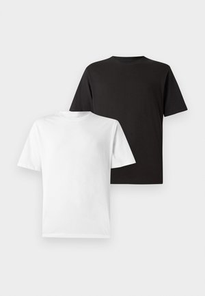 Jeune homme debout portant un t-shirt blanc uni à manches courtes, un pantalon noir et des baskets beiges sur fond blanc.