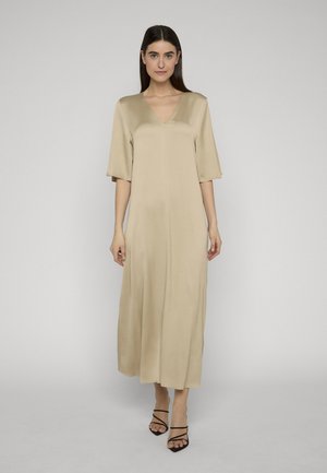 Kvinde stående mod en ensfarvet baggrund iført en beige midi-kjole med V-udskæring, halvlange ærmer og sorte højhælede sandaler med stropper.