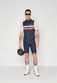 Donkerblauw fietsvest met witte en rode strepen, gedragen over een lichtgekleurde wielertrui. Navy korte broek en witte fietsschoenen maken de outfit compleet.