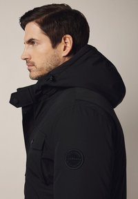 Czarna, ocieplana kurtka z kapturem, z okrągłą naszywką z logo na rękawie oraz teksturowanym materiałem. Zamknięcie z przodu na guziki oraz widoczne kieszenie.