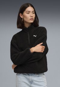 Pullover in fleece nero con colletto a zip quarti e logo bianco sul lato sinistro del petto. Texture morbida e vestibilità rilassata, abbinato a jeans azzurri chiari.