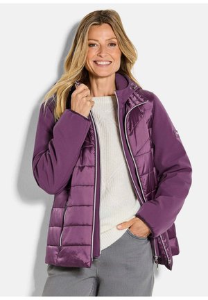 Femme souriante, portant une veste matelassée violette par-dessus un pull blanc, une main près du col et l'autre dans la poche d'un pantalon gris.