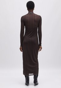Langärmliges, durchsichtiges Midikleid mit vertikalen Streifen in Dunkelbraun und Schwarz. Hat einen hohen Kragen und einen seitlichen Schlitz. Kombiniert mit schwarzen spitzen Stiefeln.