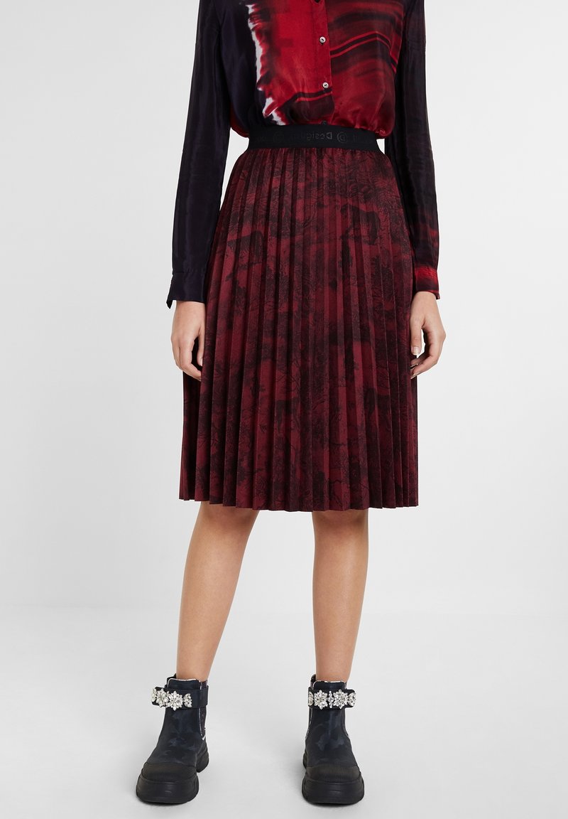 Femme porte une jupe plissée rouge et noire, un chemisier rouge à manches longues et des bottines noires avec des sangles décoratives ornées de bijoux.