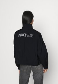 Nike Air-jacka, svart, med hög krage, oversized passform, har bakått broderi av "NIKE AIR" i vitt, slät textur.