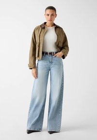 Olive green cropped jacket with brown collar, paired with light blue wide-leg jeans. Model wears a white ribbed top and black heels.