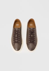Brown leather sneakers with flat dark brown laces, cream rubber soles, and embossed branding on the sides and tongue. Smooth texture.