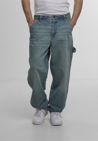 Light blue loose-fitting denim jeans with a faded texture, front pockets, and a side loop. Paired with white sneakers for contrast.