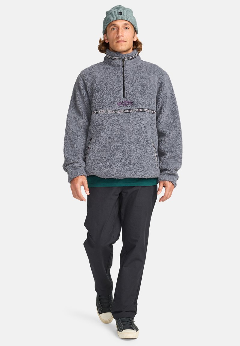 Grå fleece-pullover med halvvip, der har et mønstrede bånd over brystet. Parret med sorte bukser og en lyseblå hue.