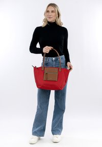 Red fabric tote bag with brown leather accents and a front zipper pocket. Features a structured shape, dual handles, and a spacious interior.