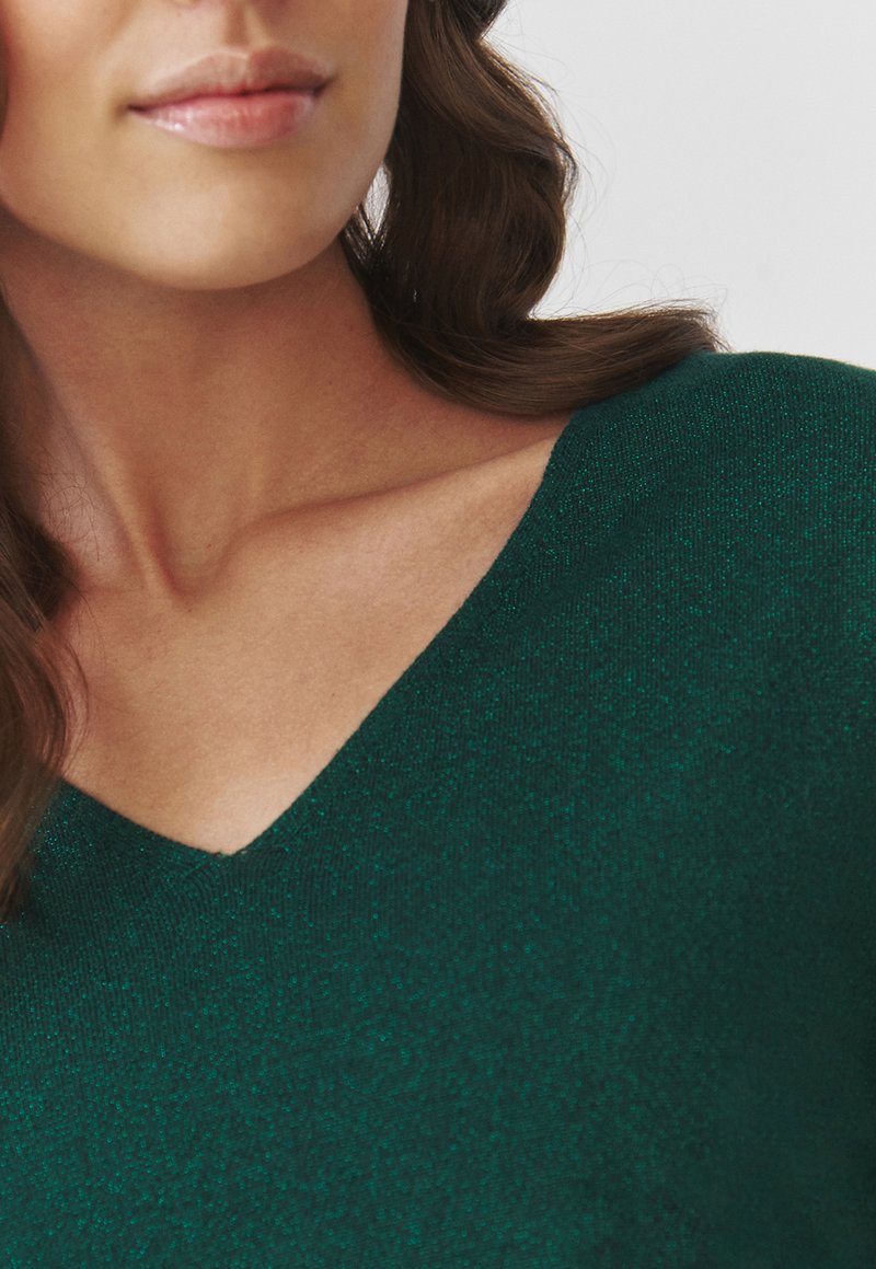 Smaragdgrünes V-Ausschnitt-Oberteil aus strukturiertem Strickstoff, mit figurnaher Passform und subtiler Glitzeroptik, die den Ausschnitt betont.