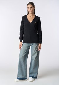 Maglione nero a collo a V con maniche lunghe, con un piccolo logo sul petto. Indossato con jeans a gamba larga azzurri e sneakers bianche.