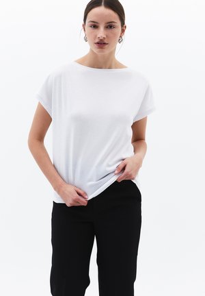Frau, die nach vorne schaut, trägt ein schlichtes weißes T-Shirt mit kurzen Ärmeln und schwarze Hosen und hält mit einer Hand den Saum ihres Shirts.