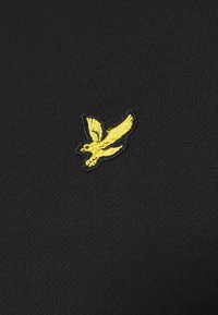 Tessuto nero con una texture a righe presenta un logo di un uccello ricamato giallo sul lato sinistro. Il design è minimalista e pulito.