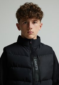 Chaleco acolchado negro con cuello alto, que presenta un cierre frontal y un panel texturizado con la marca "ZAVETTI". Tejido suave con un acabado mate.