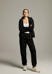 Czarna bluza z kapturem na zamek i pasujące dresowe spodnie, zestawione z białym topem na ramiączkach. Posiada luźny krój i teksturowany materiał, noszone z jasnymi sneakersami.