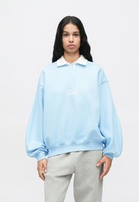 Žena s dlhými tmavými vlasmi, oblečená v svetlomodrom oversized mikinovom svetri s bielym golierom a sivých teplákoch, stojí proti bielej pozadi.