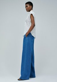 Blusa blanca de manga corta bordada combinada con jeans de mezclilla azul de pierna ancha y cintura alta. Modelo de pie, con las manos en los bolsillos.