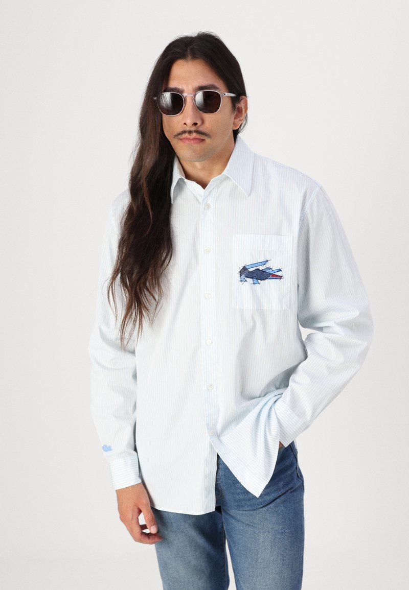 Camicia da uomo button-up in azzurro chiaro con righe bianche, caratterizzata da una tasca sul petto con un design ricamato blu e rosso. Tessuto leggero.