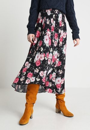 Woman wearing a black floral midi skirt, navy cable-knit sweater, and mustard suede heeled boots standing on white floor.