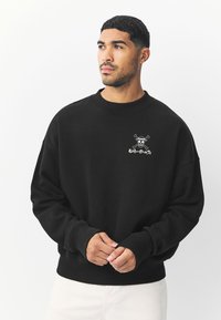Sort sweatshirt med rund hals og løs pasform. Har et grafisk design på brystet, der viser en karakter med solbriller og korslagte knogler.