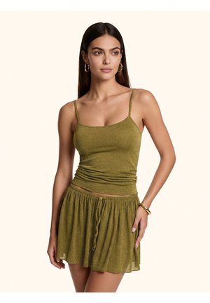 Donna con lunghi capelli scuri indossa orecchini a cerchio dorati, top verde oliva aderente e minigonna plissettata abbinata con coulisse.